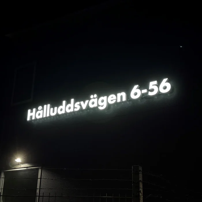 Vit LED-belyst fasadskylt med adressen "Hålluddsvägen 6–56", monterad på en mörk fasad – visar hur skyltar på fasad kan användas för att tydligt markera adress.