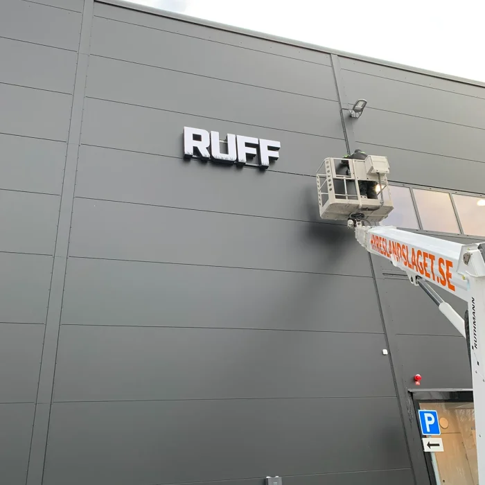 Företagsskylt i upphöjda bokstäver monteras på en grå industrifasad med hjälp av skylift – ett bra exempel på fasadskyltar för företag med robust design och tydlig placering.