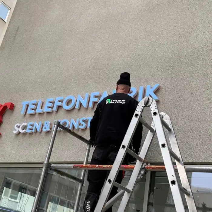 Skyltmontage Stockholm - En arbetare installerar en fasadskylt med texten "TELEFONFABRIKEN" på en byggnad i Stockholm.