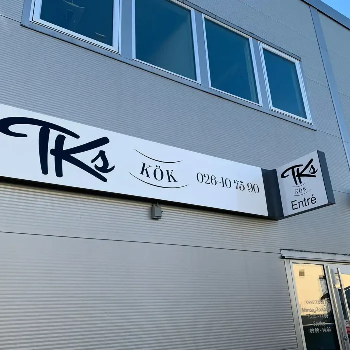 En stilren vit plåtskylt med svart text för "Tks Kök", monterad på en industribyggnad. Skylten innehåller företagets namn, telefonnummer och en mindre entréskylt.